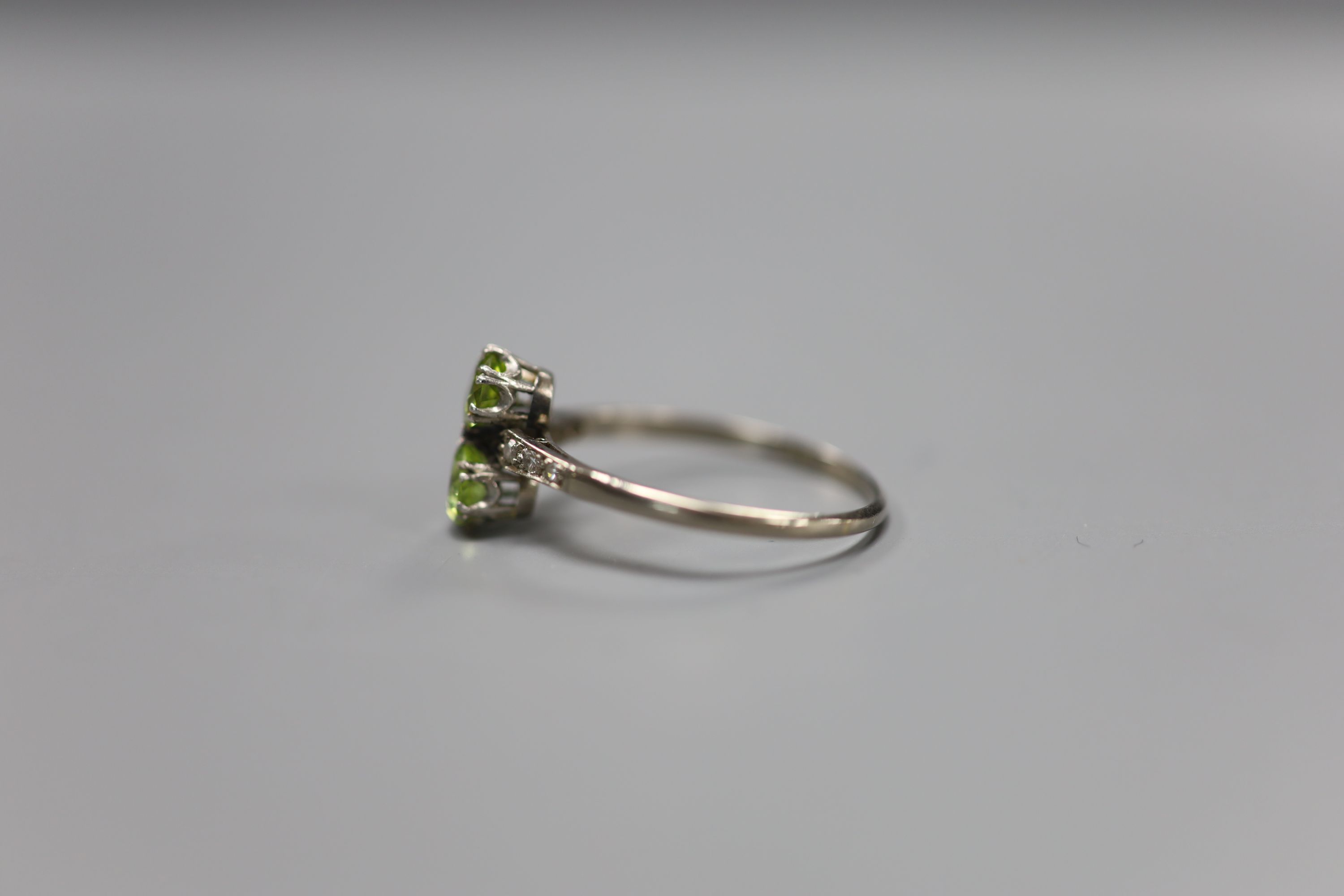 An 18ct and plat, two stone peridot crossover ring, with diamond set shoulders, size W, gross 2.7 grams.